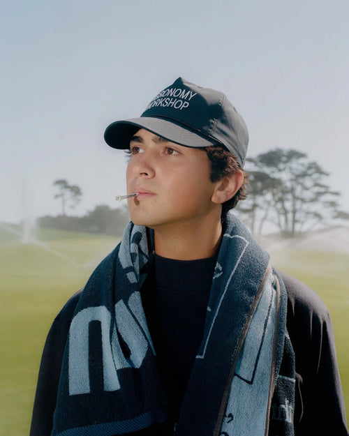 A man wearing a dark navy and gray baseball cap with 'AGRONOMY WORKSHOP' embroidered on it, a dark navy scarf with light blue lettering, and a black t-shirt, standing on a golf course with sprinklers in the background.