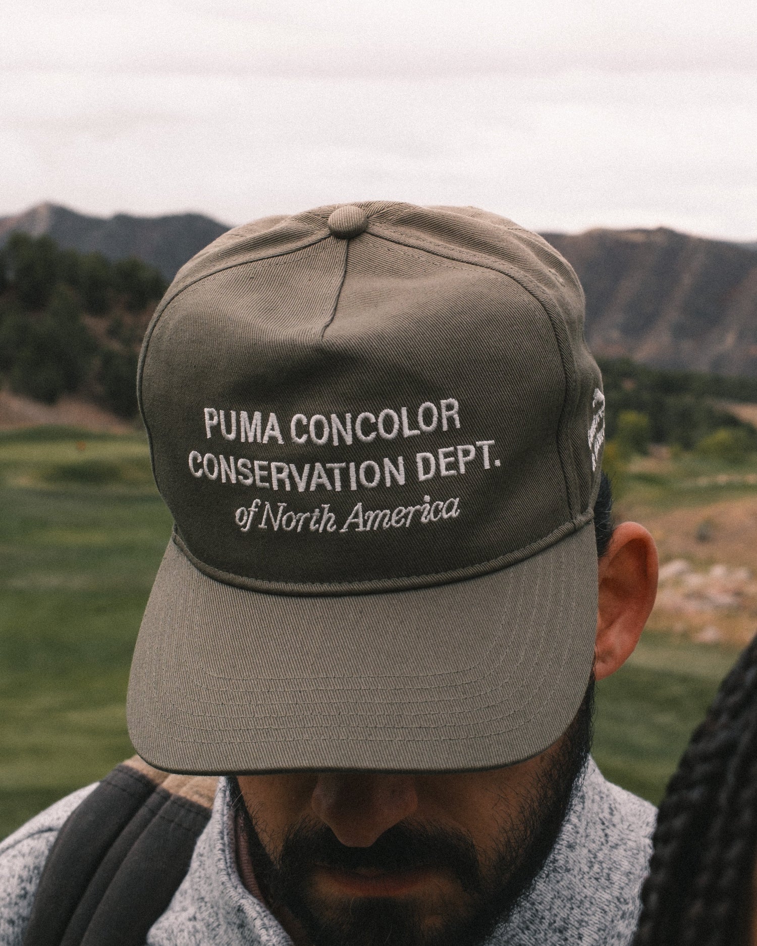 Person wearing a cap with 'Puma Concolor Conservation Dept. of North America' text in a natural setting.