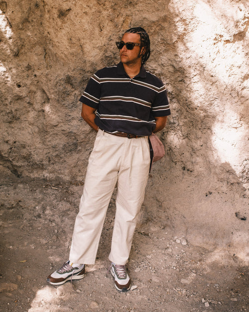 Man wearing a striped shirt and white pants standing against a textured wall.