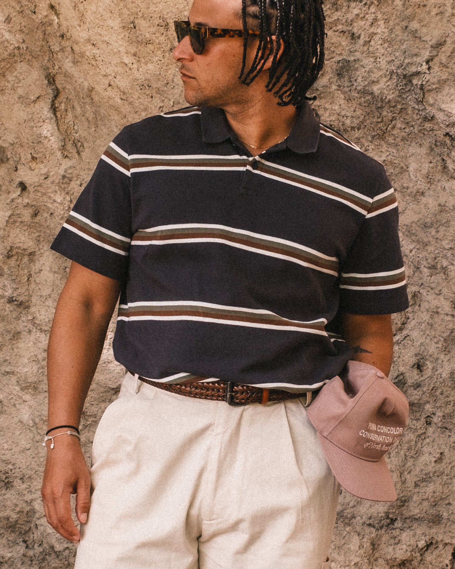 Man wearing a striped polo shirt and sunglasses, standing against a textured wall.