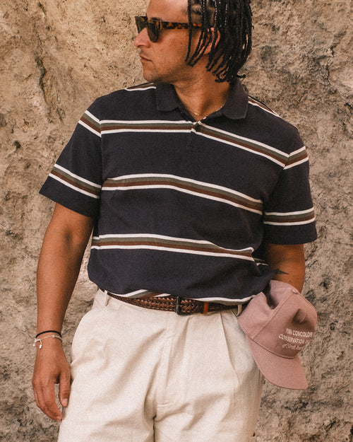 Man wearing a striped polo shirt and sunglasses, standing against a textured wall.