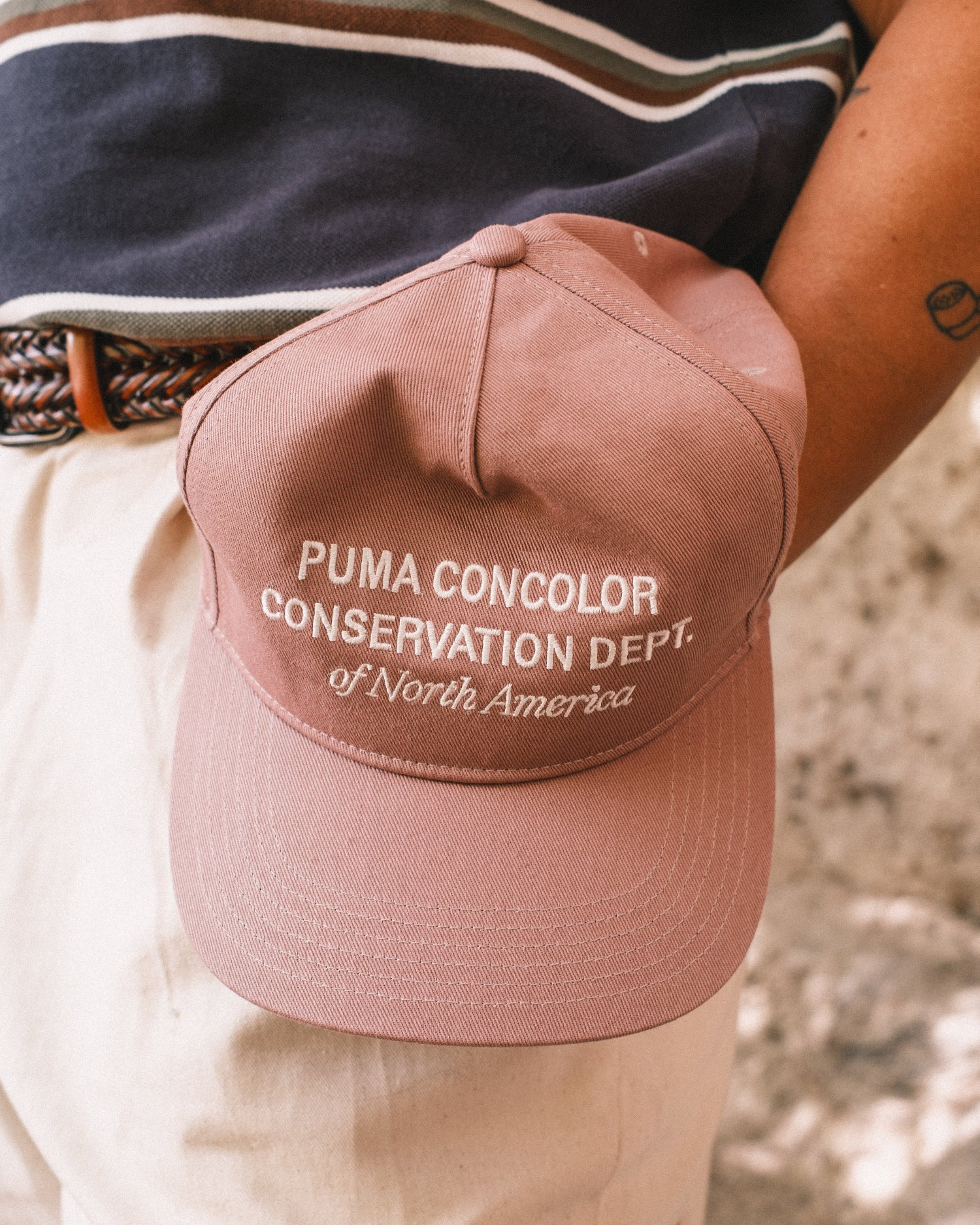 Brown cap with text held by a person wearing a striped shirt and beige pants.