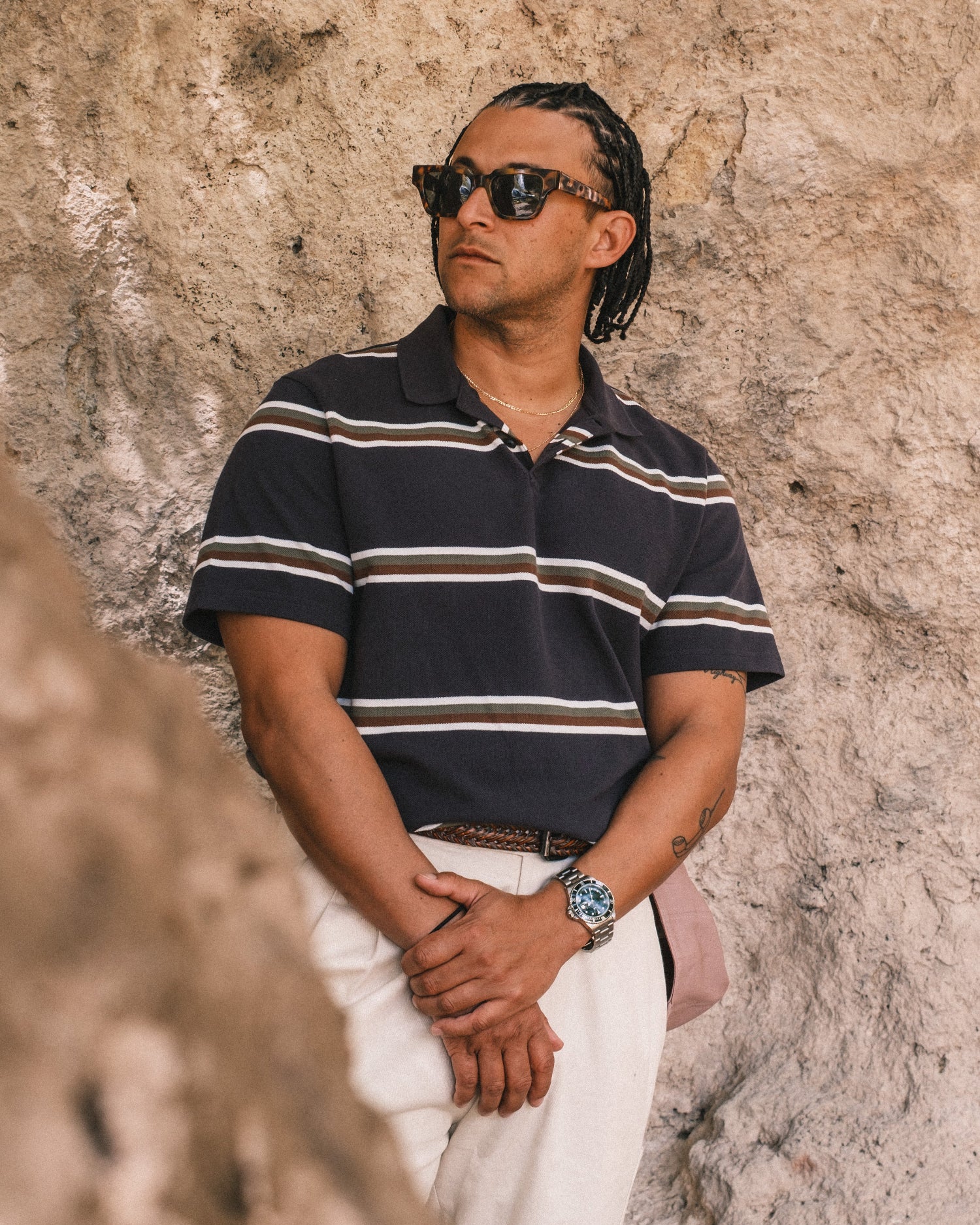 Man wearing a striped polo shirt and sunglasses against a textured wall.