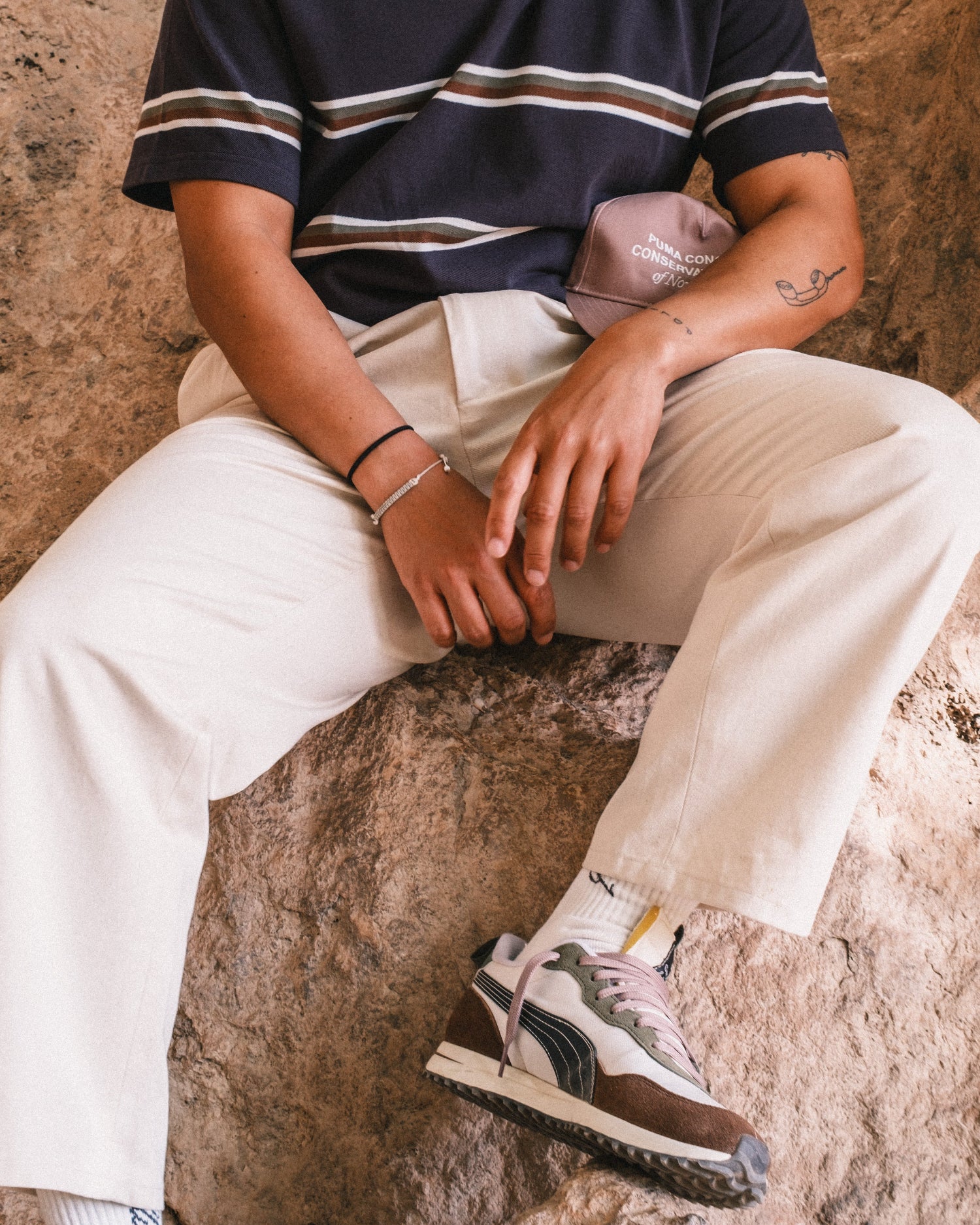 Person wearing a striped shirt, light pants, and sneakers sitting on a stone surface.