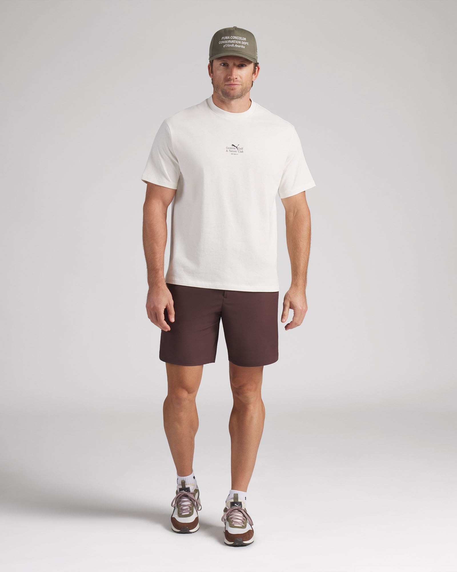 Man wearing a white t-shirt, brown shorts, and a cap on a plain background