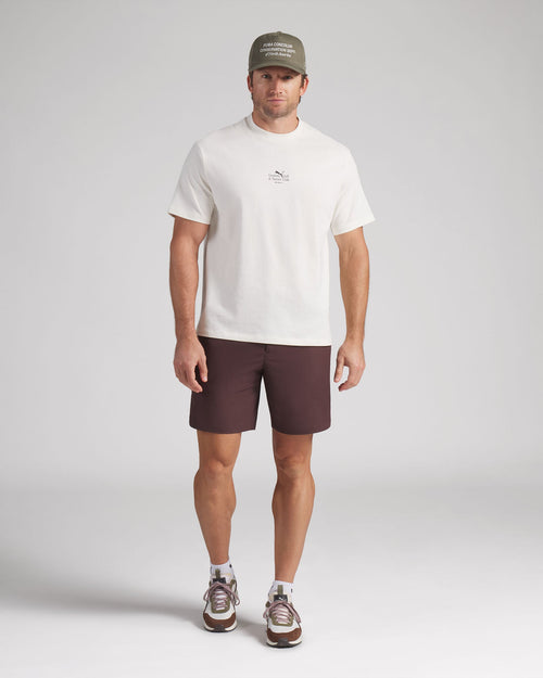 Man wearing a white t-shirt, brown shorts, and a cap on a plain background