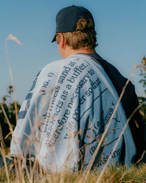 A man wearing a black baseball cap sits outdoors in a grassy field, viewed from behind. He has a light blue knit blanket draped over his shoulders, featuring black printed text and diagrams.